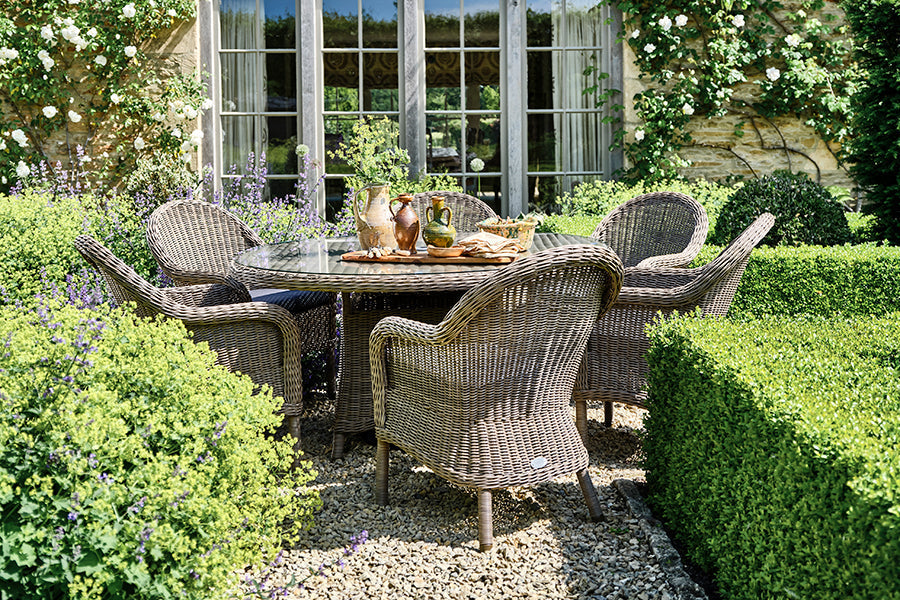 Luxury rattan garden dining set (round dining table with six dining armchairs) sits in a sunny garden surrounded by hedges.